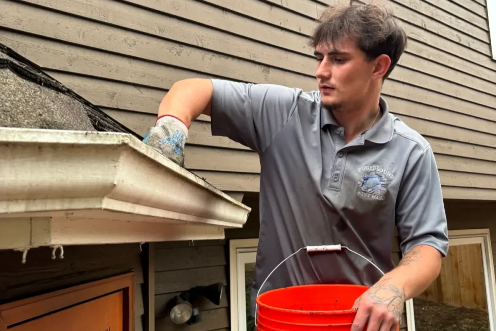 "Person cleaning gutters with a glove and placing debris into a bucket near a wooden house