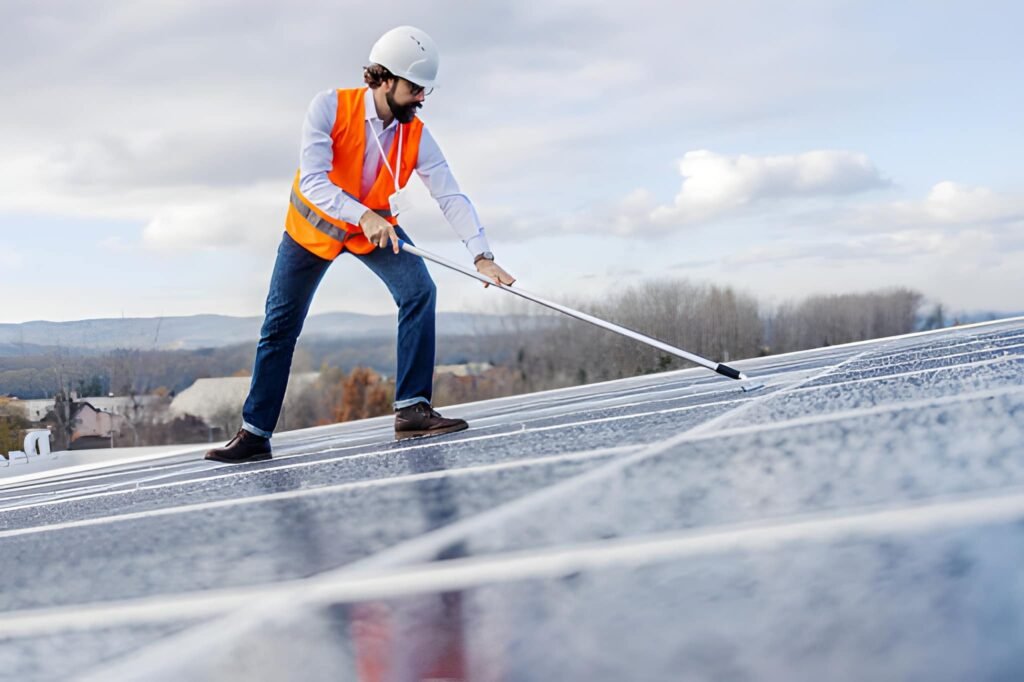 Renewable energy technician in hard hat and safety vest clears water from rooftop solar panels with a broom